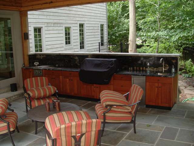 Outdoor kitchen on sheltered stone patio with seating area in the foreground that was designed by a Landscape Architect.